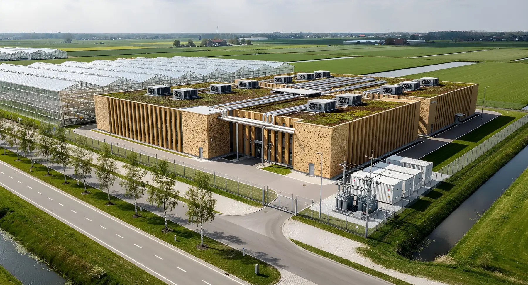 Aerial view of a compact data centre development with a tree-lined connecting road running to its entrance, a fenced substation with battery containers, heat pipes crossing to a block of commercial glasshouses along one side, and open polder fields beyond